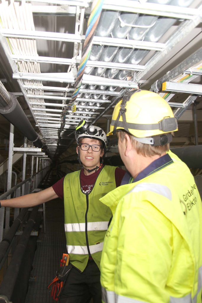 Elektrikere fra Årnes Elektro jobber med kabler og elektrisk utstyr i en teknisk korridor, utstyrt med sikkerhetsutstyr som hjelmer og refleksvester.